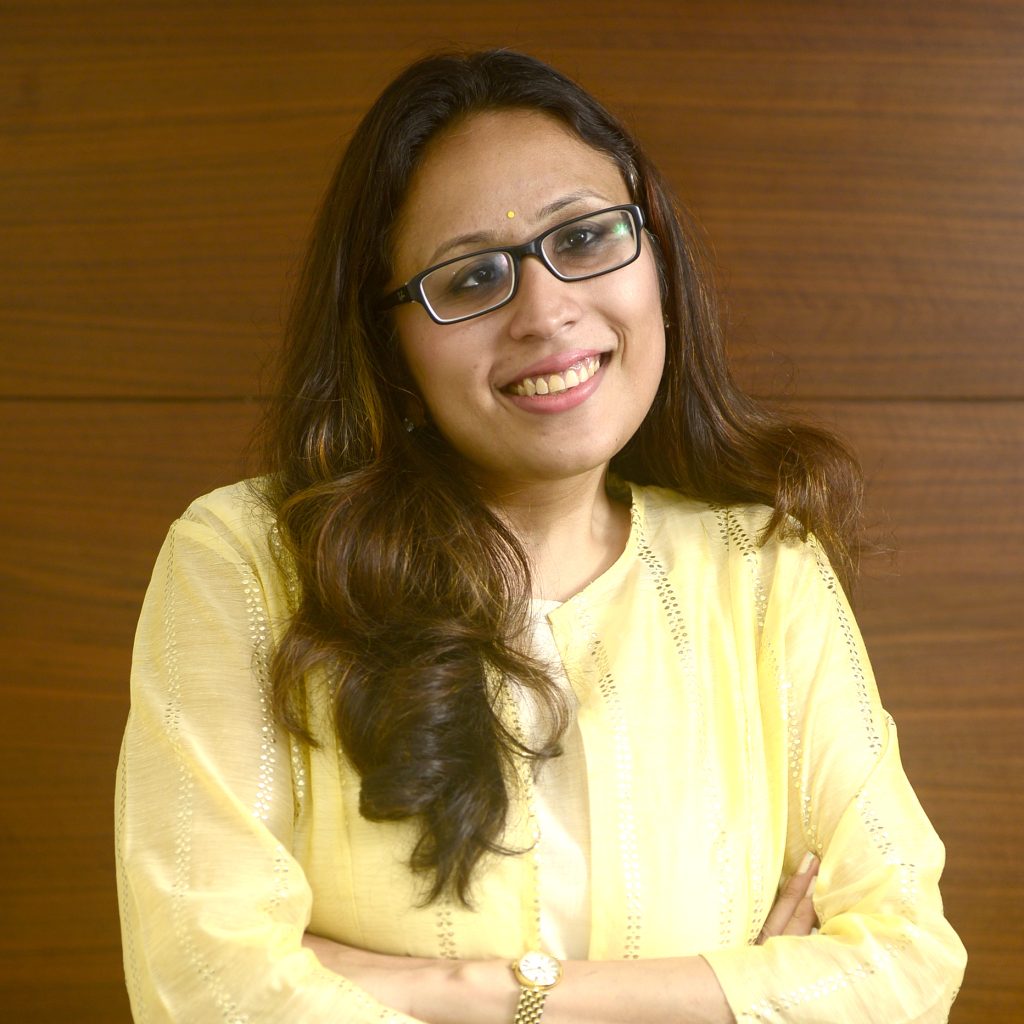 Radhika Gupta in glasses and a yellow kurta, smiling with her arms folded