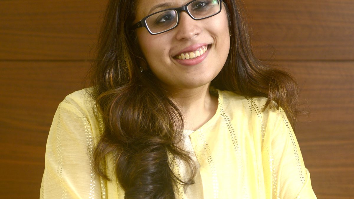 Radhika Gupta in glasses and a yellow kurta, smiling with her arms folded