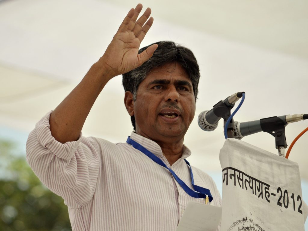 Rajagopal P. V. raising his hand as he speaks into microphones at an open-air rally