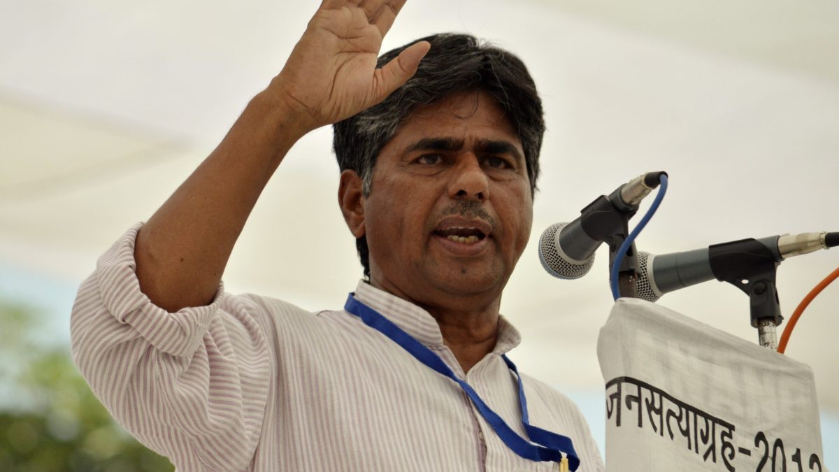 Rajagopal P. V. raising his hand as he speaks into microphones at an open-air rally