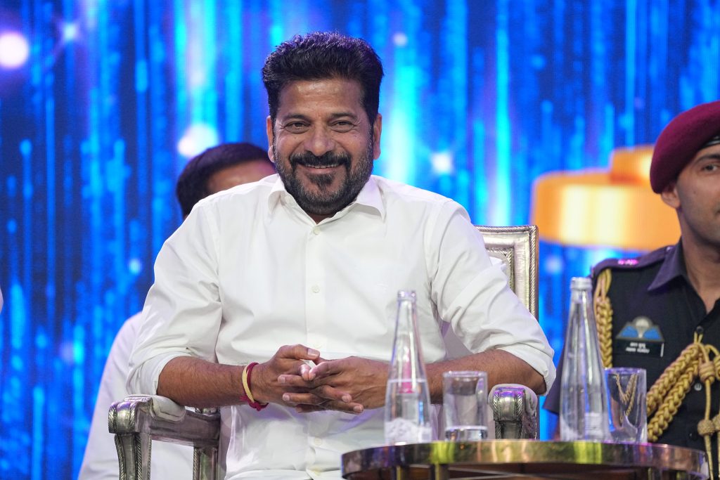 Revanth Reddy in a white shirt seated and smiling, with a blue backdrop behind him