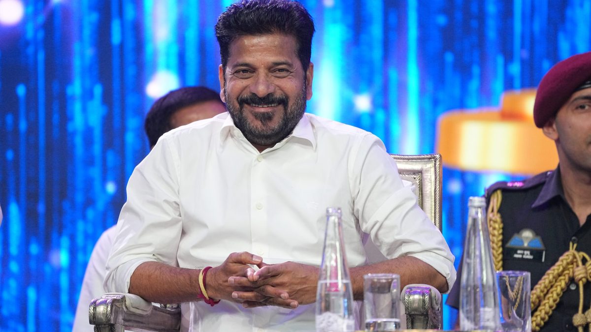 Revanth Reddy in a white shirt seated and smiling, with a blue backdrop behind him