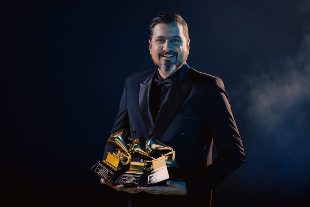 Ricky Kej in a dark suit holding three Grammy trophies against a dark background