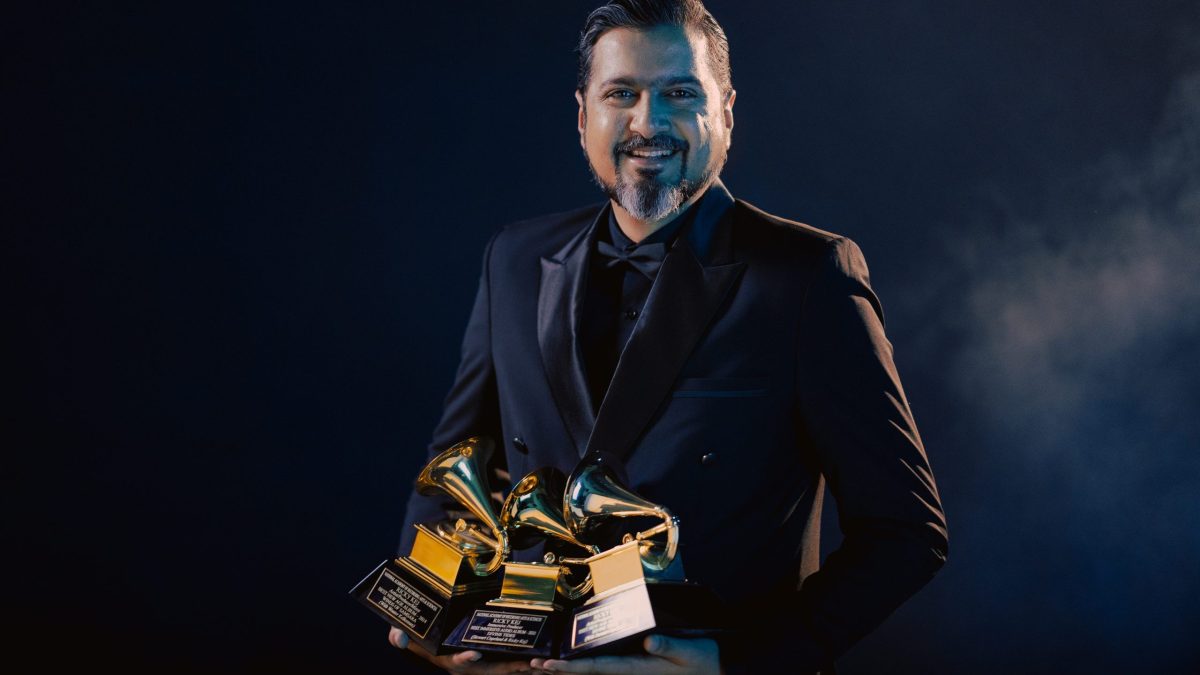 Ricky Kej in a dark suit holding three Grammy trophies against a dark background