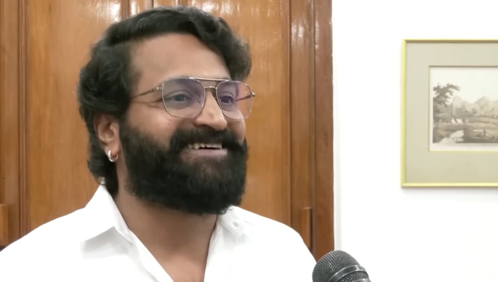 Rishab Shetty with a full beard, in a white shirt, smiling