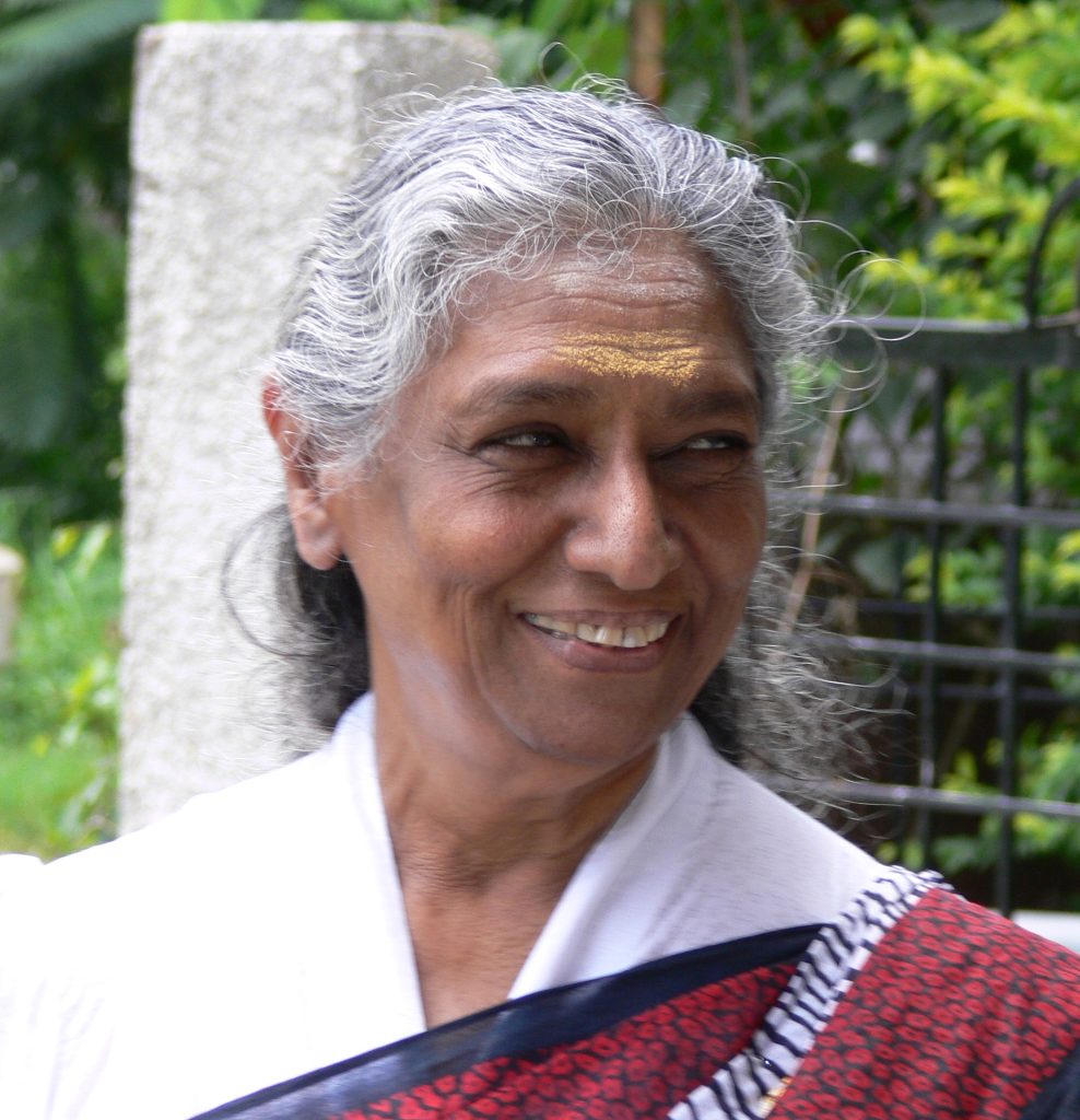 S. Janaki with white hair, smiling, in a white sari with a red border