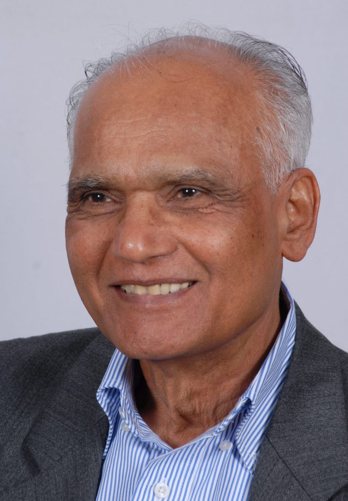S. L. Bhyrappa with white hair, smiling, in a dark jacket and striped shirt