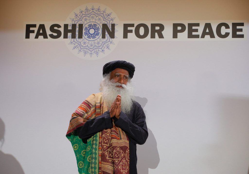 Sadhguru with a long white beard and turban greeting with folded hands under a Fashion for Peace sign