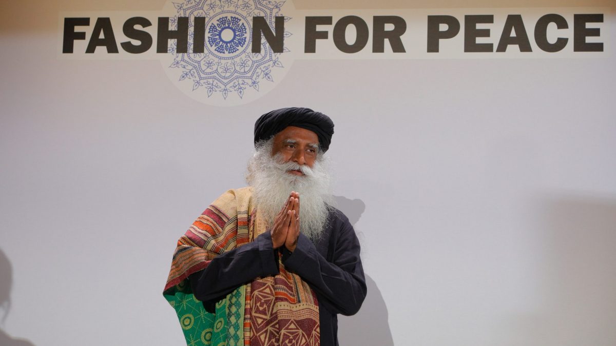 Sadhguru with a long white beard and turban greeting with folded hands under a Fashion for Peace sign