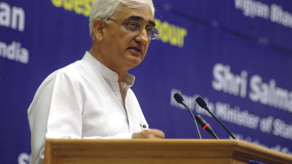 Salman Khurshid in a white kurta and glasses speaking at a lectern