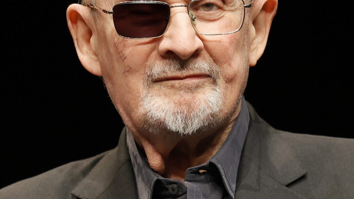 Salman Rushdie wearing glasses with one darkened lens, speaking on stage