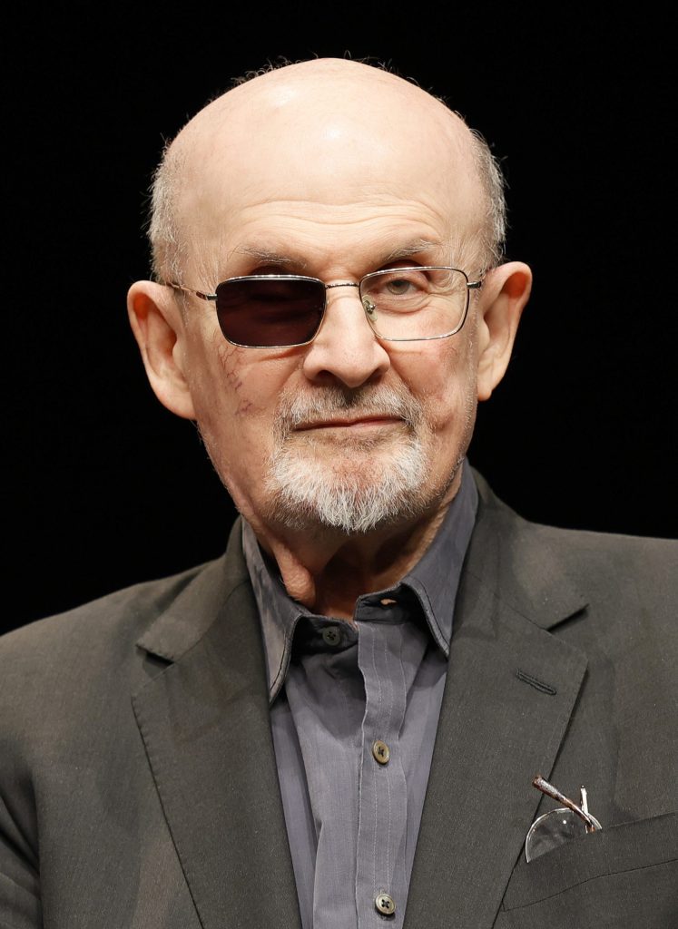 Salman Rushdie wearing glasses with one darkened lens, speaking on stage