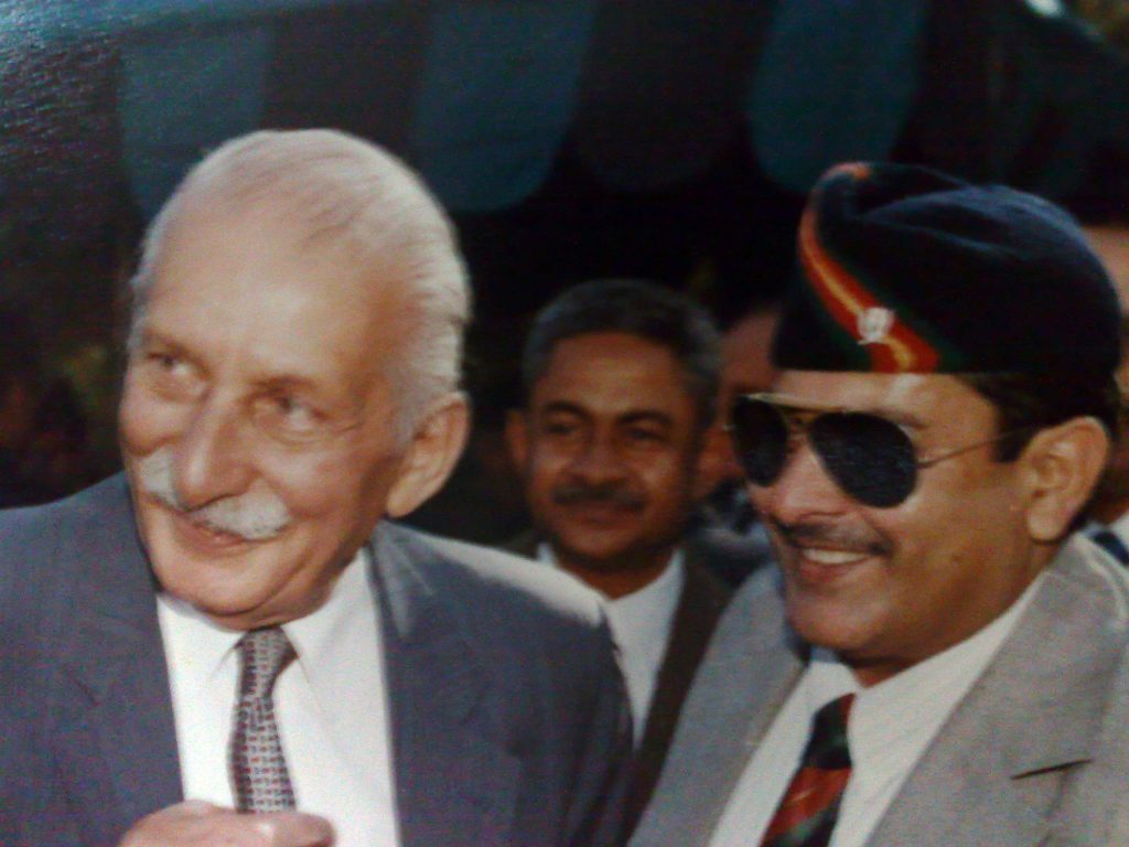 Close-up of Sam Manekshaw with a white moustache, smiling in a suit, with army officers behind him