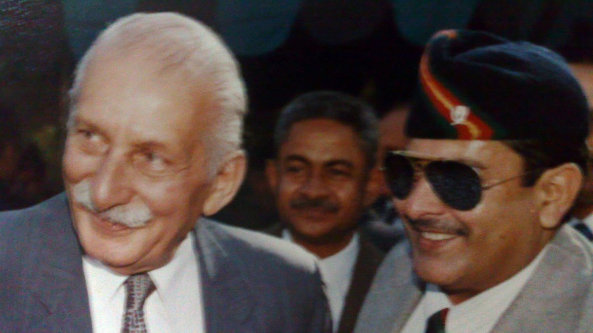 Close-up of Sam Manekshaw with a white moustache, smiling in a suit, with army officers behind him