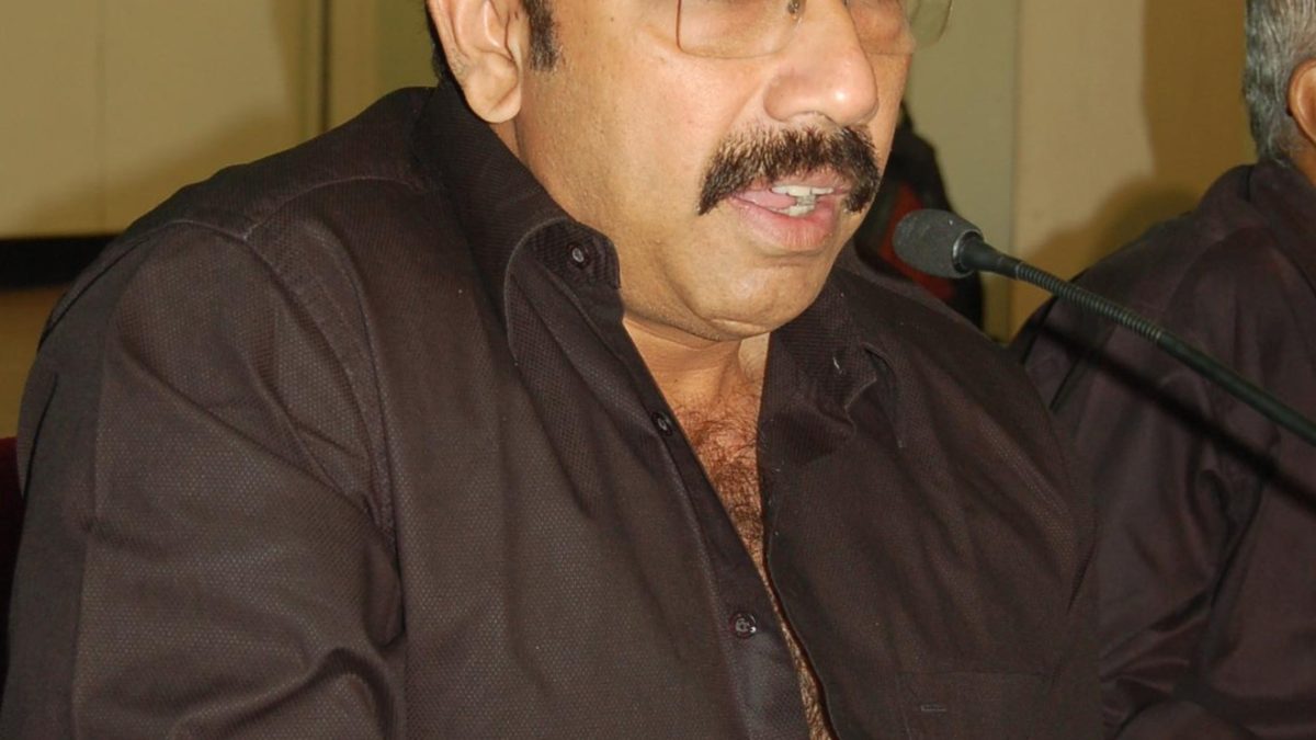 Sathyaraj in glasses and a dark shirt speaking at a table with microphones
