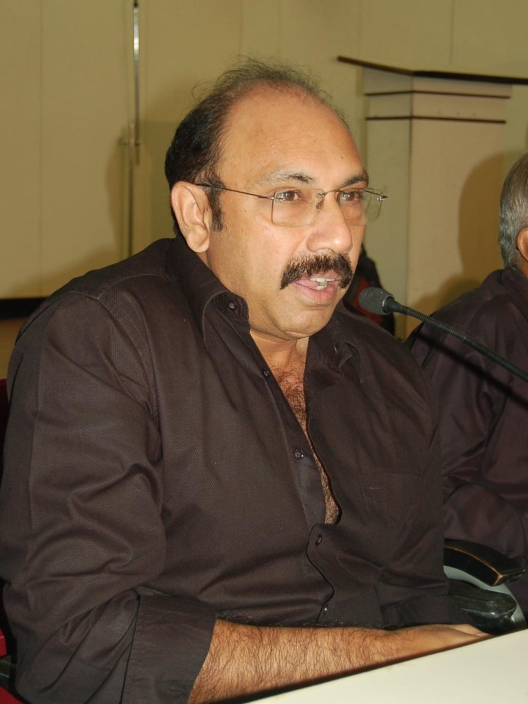 Sathyaraj in glasses and a dark shirt speaking at a table with microphones