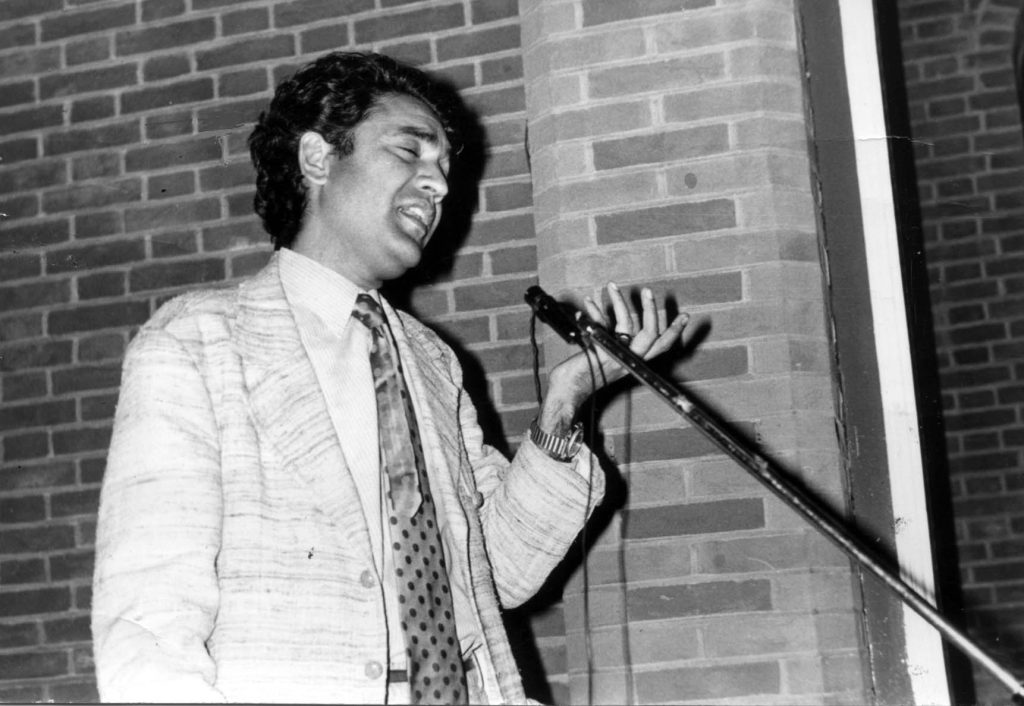 A black-and-white photograph of Shiv Kumar Batalvi in a patterned jacket reciting into a microphone