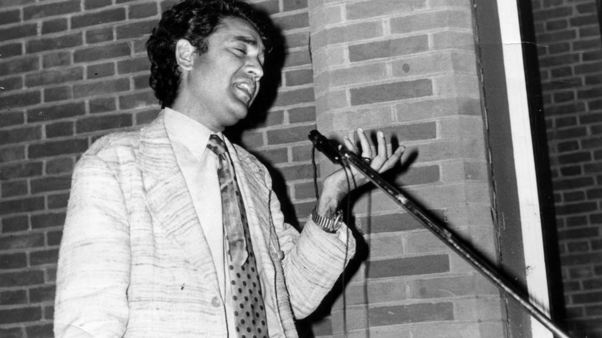 A black-and-white photograph of Shiv Kumar Batalvi in a patterned jacket reciting into a microphone