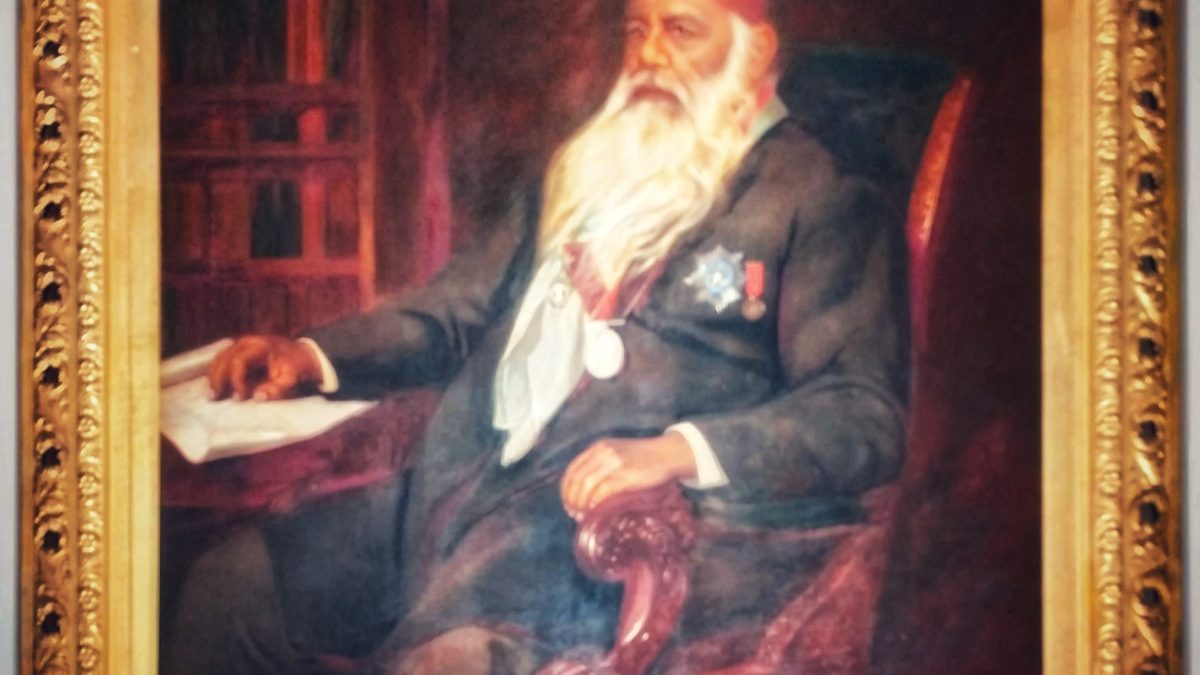 Framed oil portrait of Sir Syed Ahmad Khan with a long white beard and red fez, seated at a desk