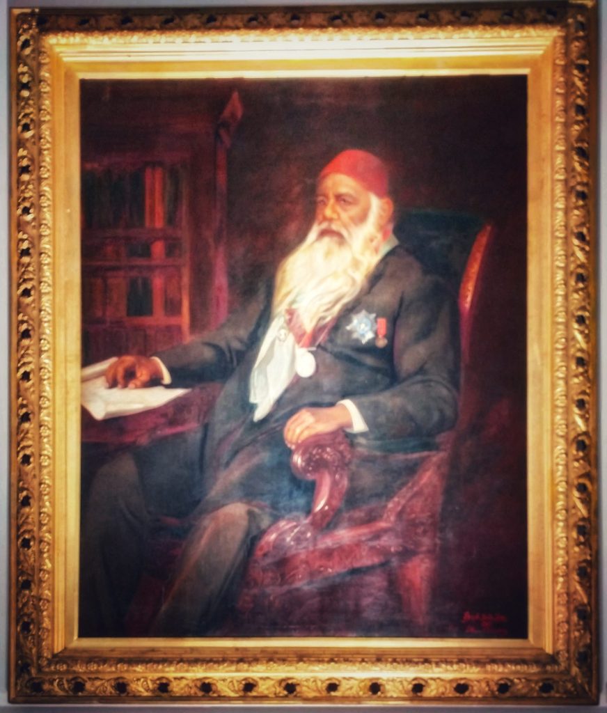 Framed oil portrait of Sir Syed Ahmad Khan with a long white beard and red fez, seated at a desk