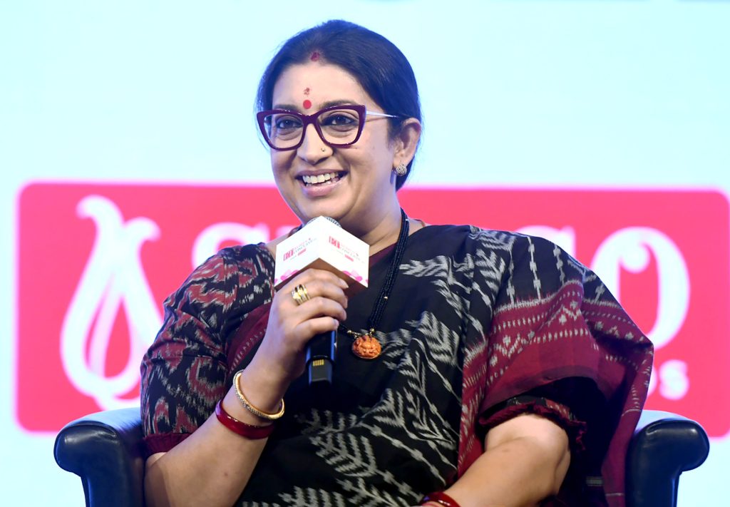 Smriti Irani in glasses and a dark sari speaking into a microphone on stage