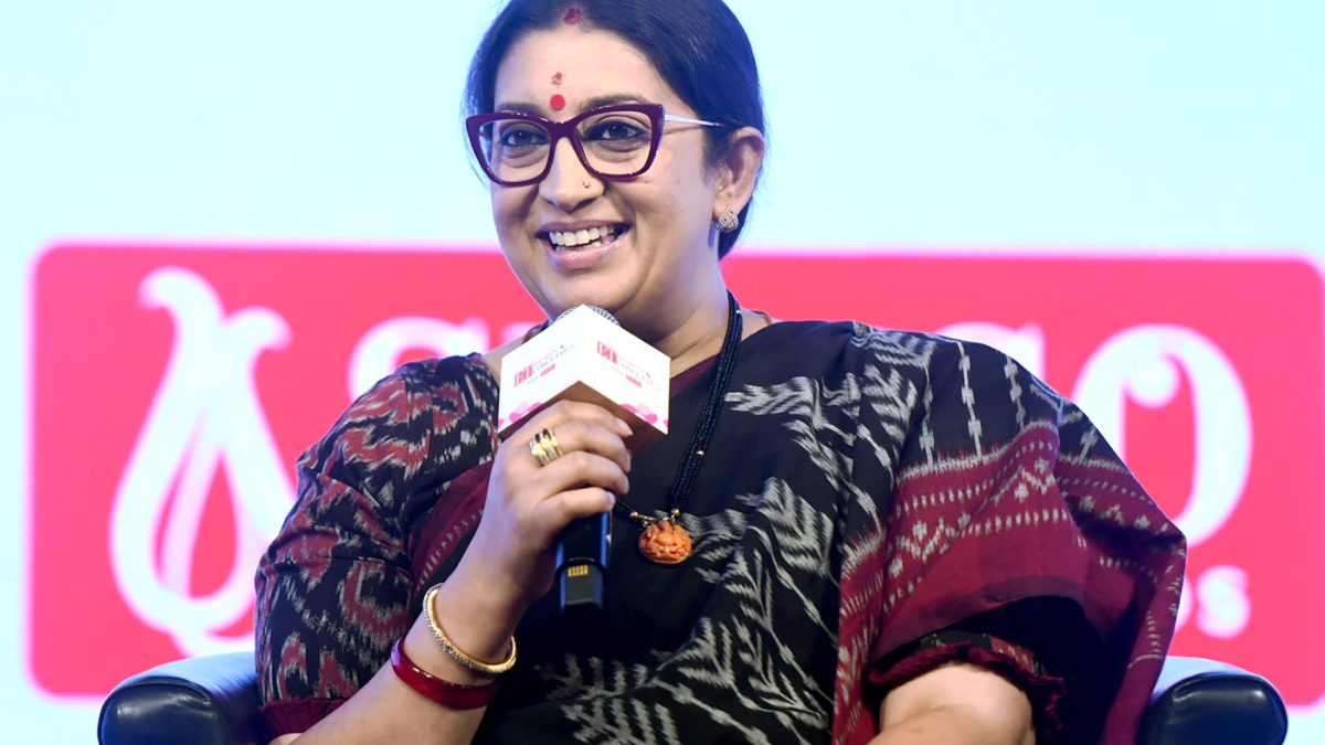 Smriti Irani in glasses and a dark sari speaking into a microphone on stage
