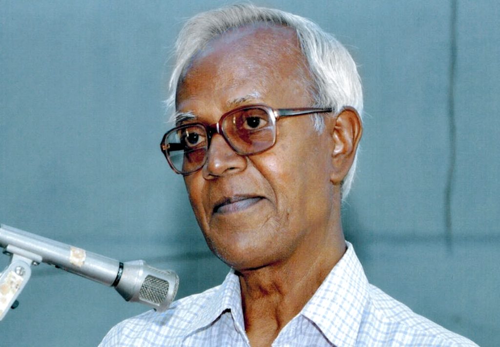 Stan Swamy with white hair and glasses speaking into a microphone