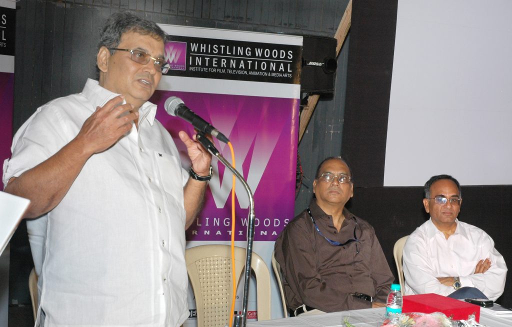 Subhash Ghai in a white shirt speaking into a microphone, with two men seated behind him