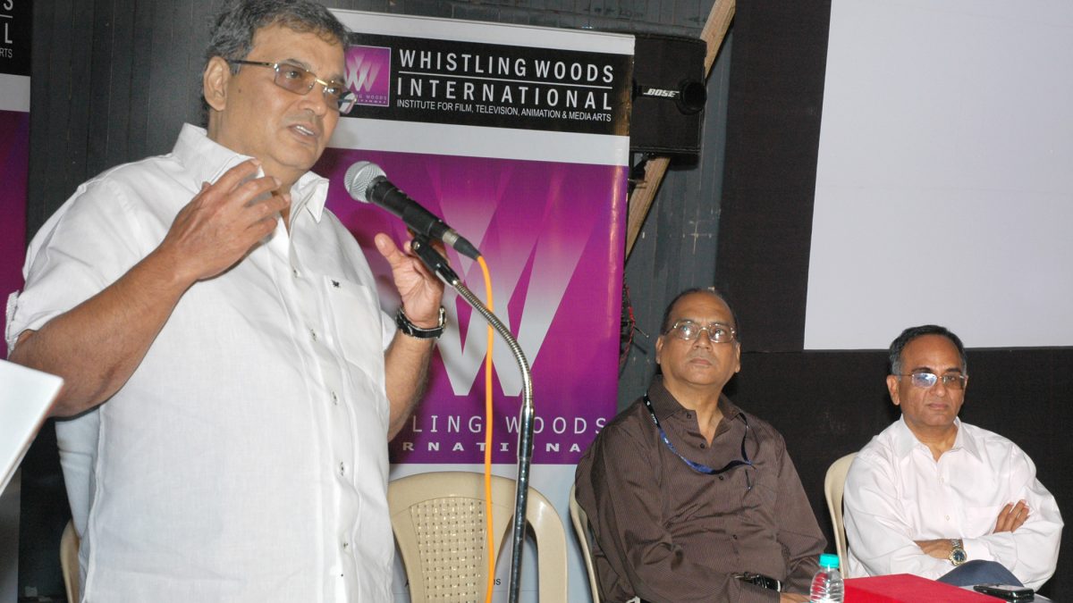 Subhash Ghai in a white shirt speaking into a microphone, with two men seated behind him