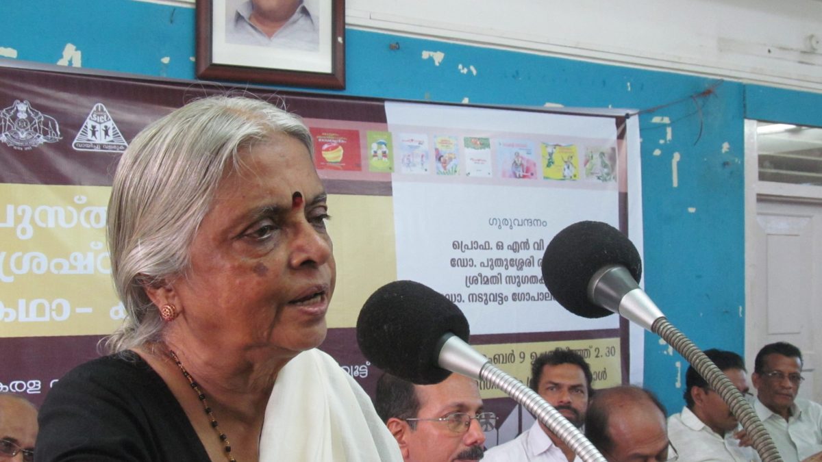 Sugathakumari with grey hair speaking into microphones at a meeting