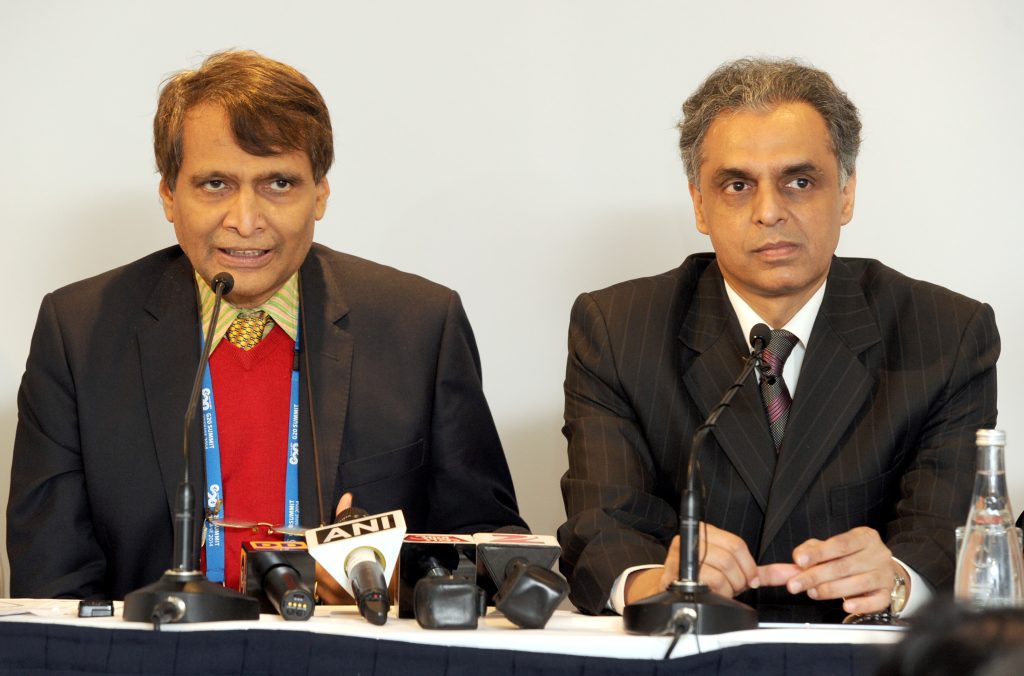 Suresh Prabhu in glasses, a dark jacket and red scarf speaking at a press conference beside an official