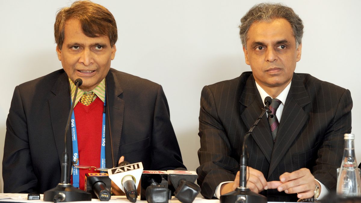 Suresh Prabhu in glasses, a dark jacket and red scarf speaking at a press conference beside an official