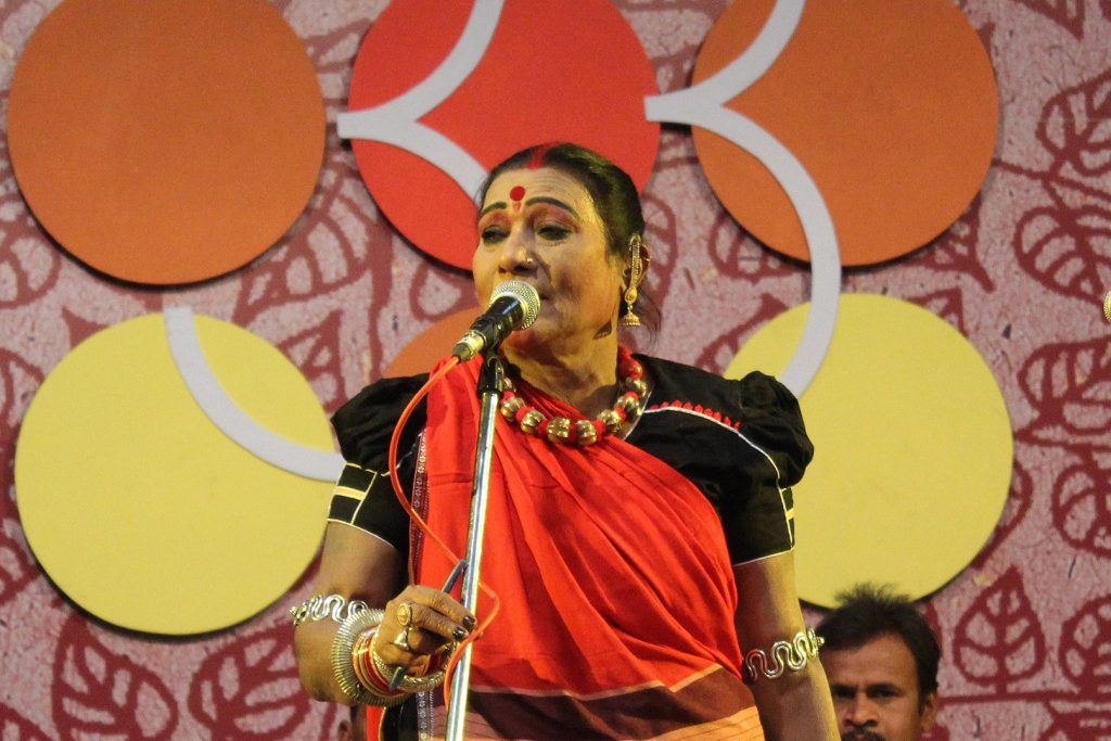 Teejan Bai in a red sari and traditional jewellery singing into a microphone