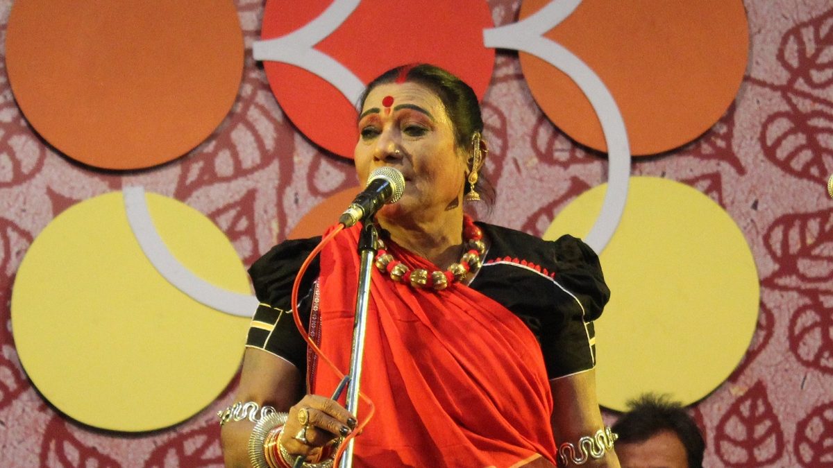 Teejan Bai in a red sari and traditional jewellery singing into a microphone
