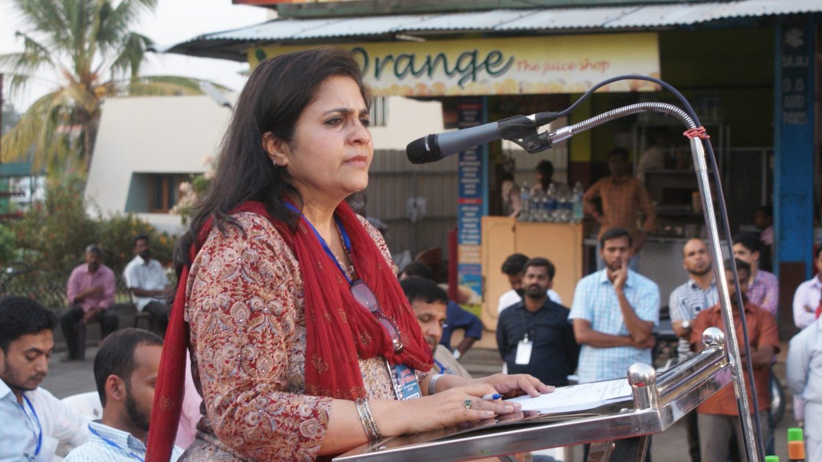 Teesta Setalvad in a red dupatta speaking at a lectern with microphones outdoors