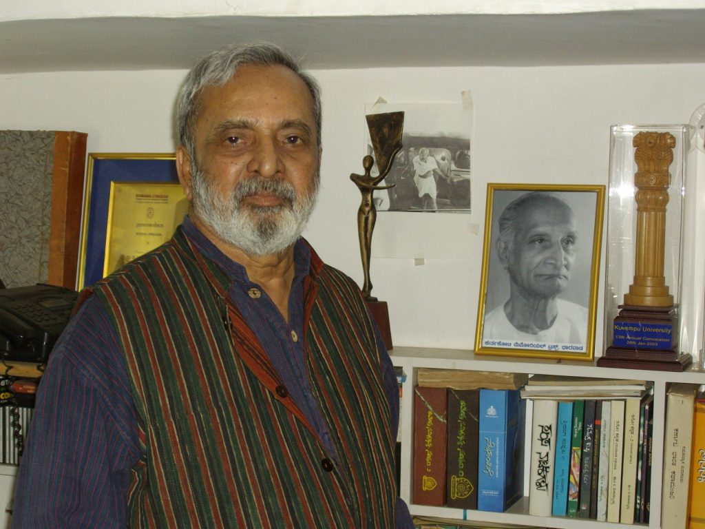 U. R. Ananthamurthy with a grey beard, in a striped waistcoat, standing before bookshelves