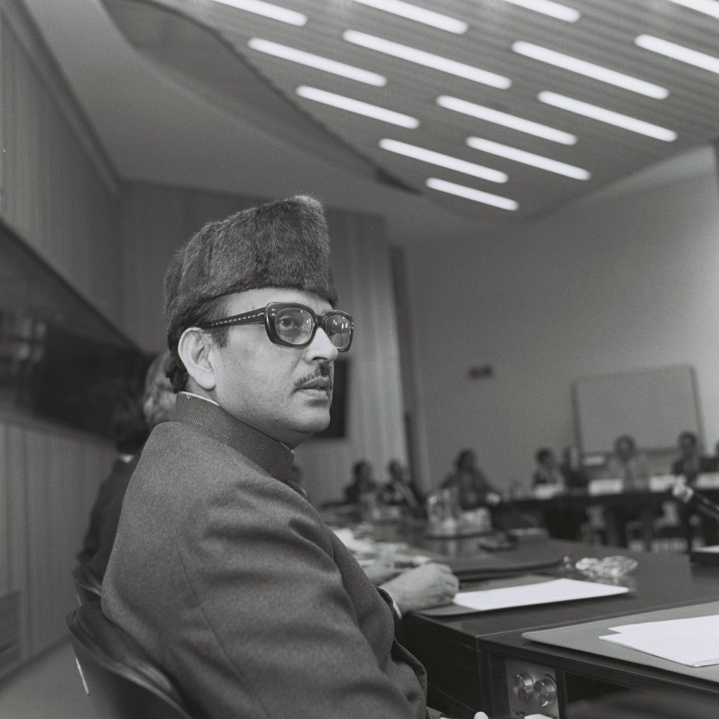 Black-and-white photograph of V. P. Singh in a fur cap and glasses seated at a conference table