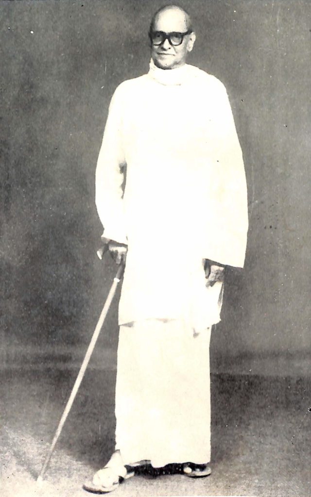 Full-length black-and-white photograph of Vallathol in a white shirt and mundu, holding a walking stick