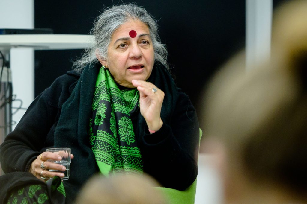Vandana Shiva with grey hair, a red bindi and green scarf, speaking at an event