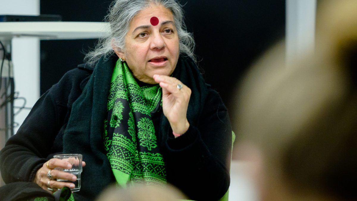 Vandana Shiva with grey hair, a red bindi and green scarf, speaking at an event