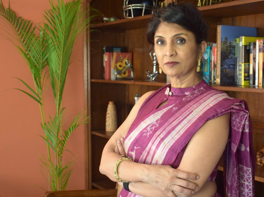 Vani Kola in a striped sari standing with arms folded in front of bookshelves