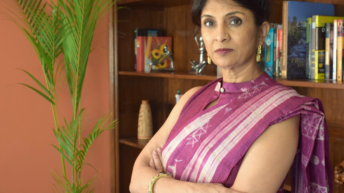 Vani Kola in a striped sari standing with arms folded in front of bookshelves