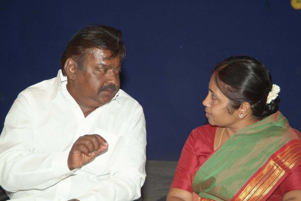 Vijayakanth in a white shirt seated at a function, talking with a woman beside him