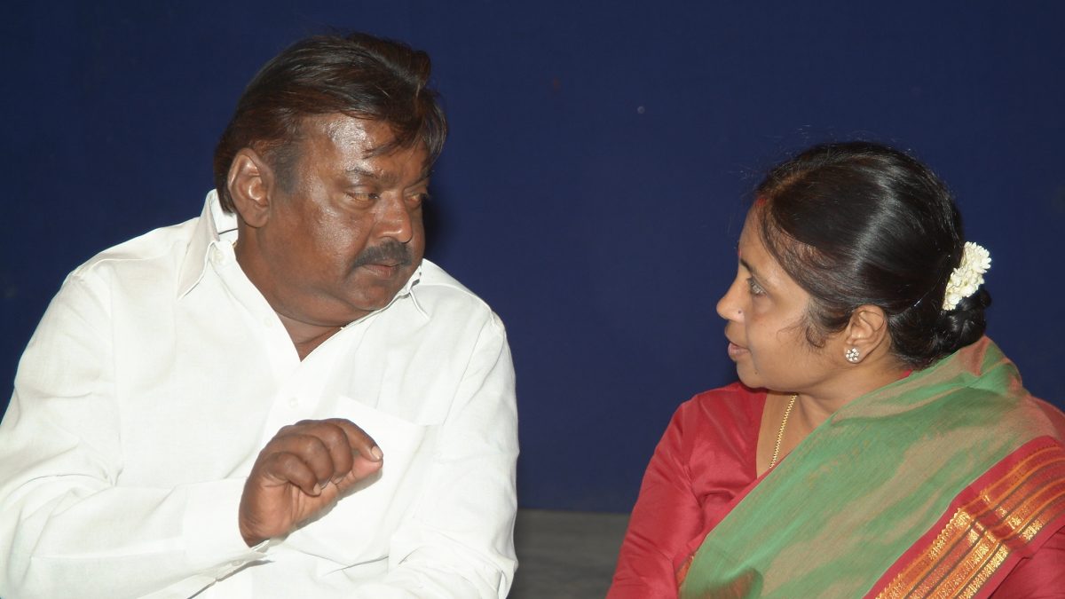 Vijayakanth in a white shirt seated at a function, talking with a woman beside him