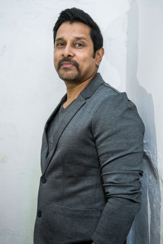 Vikram with a moustache, in a grey jacket, standing with his hands in his pockets