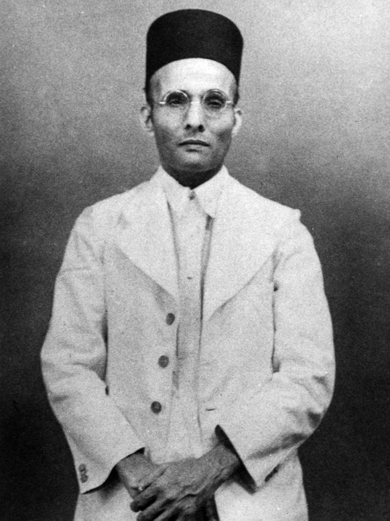 A black-and-white photograph of the young Savarkar standing, in a cap and glasses