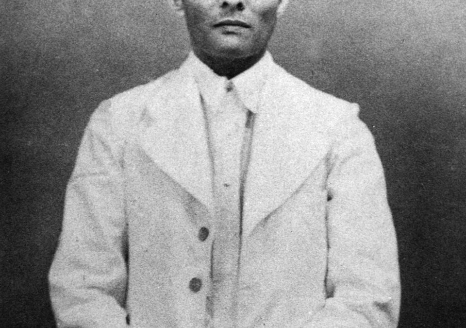 A black-and-white photograph of the young Savarkar standing, in a cap and glasses