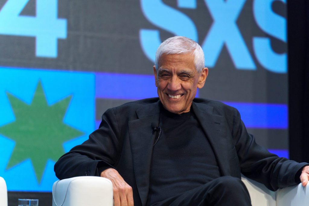 Vinod Khosla with white hair in a dark jacket seated on stage, smiling