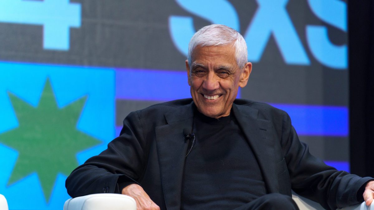 Vinod Khosla with white hair in a dark jacket seated on stage, smiling