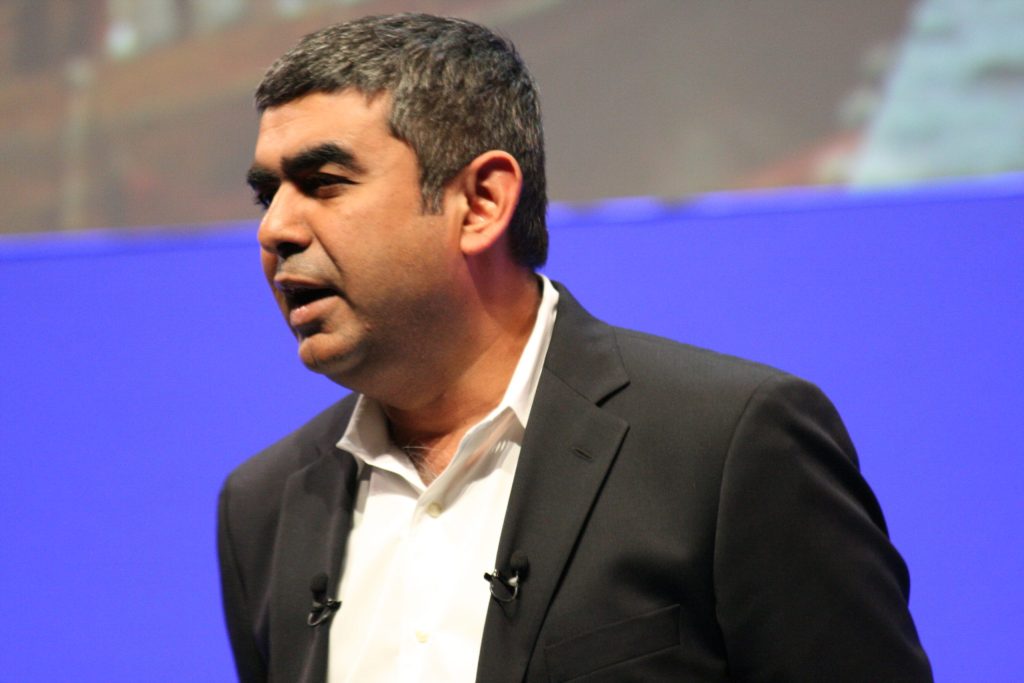 Vishal Sikka in a dark jacket speaking on stage against a blue background